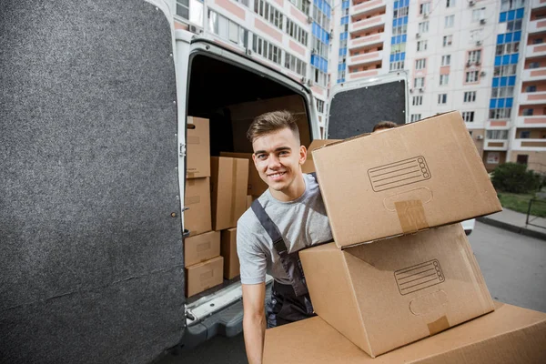 Dos jóvenes y guapos trabajadores sonrientes con uniformes están descargando la camioneta llena de cajas. El bloque de pisos está en el fondo. Movimiento de casa, servicio de mudanza — Foto de Stock