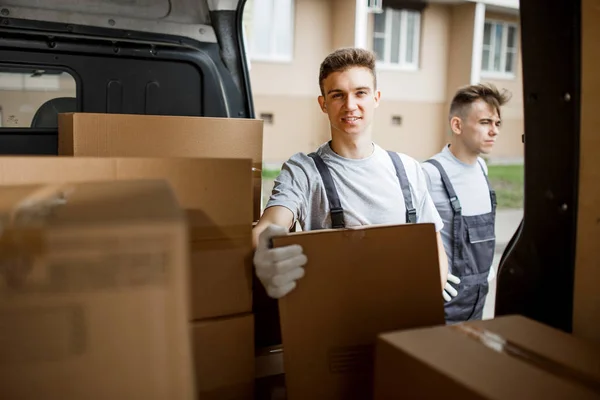 To unge, kjekke arbeidere i uniform står ved bilen full av esker. Flytting av hus, flyttetjeneste – stockfoto