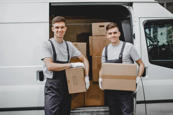 Двое молодых красивых улыбающихся рабочих в форме стоят рядом с фургоном, полным коробок. Переезд, перевозка — стоковое фото