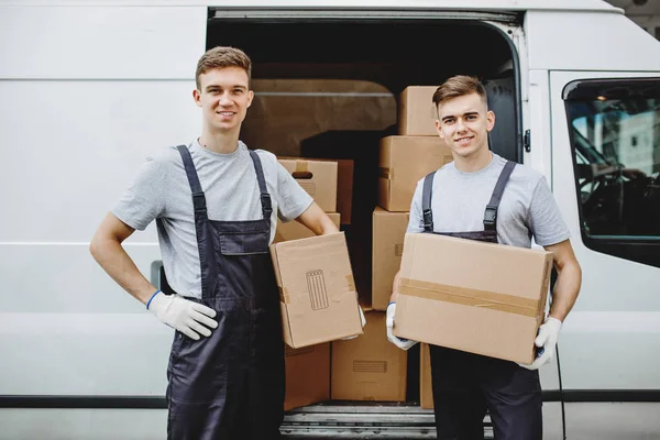 Двое молодых красивых улыбающихся рабочих в форме стоят рядом с фургоном, полным коробок. Переезд, перевозка — стоковое фото