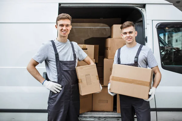 Dos jóvenes y guapos trabajadores sonrientes con uniformes están de pie junto a la furgoneta llena de cajas. Movimiento de casa, servicio de mudanza — Foto de Stock