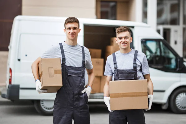 Dos jóvenes y guapos trabajadores sonrientes con uniformes están de pie junto a la furgoneta llena de cajas con cajas en las manos. Movimiento de casa, servicio de mudanza — Foto de Stock