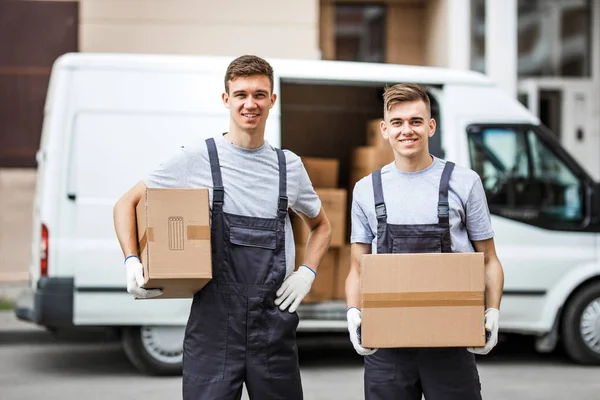 Dos jóvenes y guapos trabajadores sonrientes con uniformes están de pie junto a la furgoneta llena de cajas con cajas en las manos. Movimiento de casa, servicio de mudanza — Foto de Stock