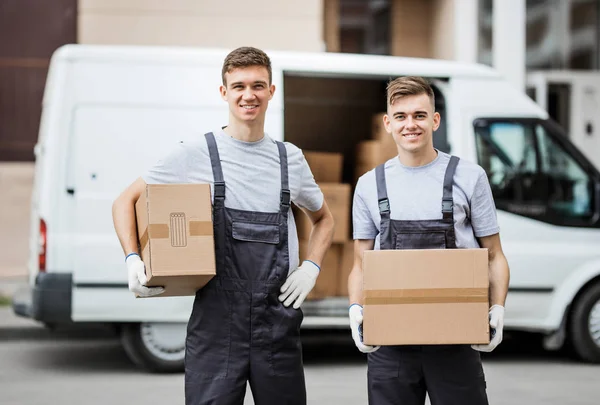 Dos jóvenes y guapos trabajadores sonrientes con uniformes están de pie frente a la camioneta llena de cajas con cajas en las manos. Movimiento de casa, servicio de mudanza — Foto de Stock