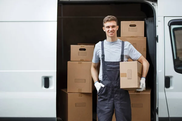 Seorang pemuda tampan tersenyum pekerja mengenakan seragam berdiri di samping van penuh kotak memegang kotak di tangannya. Rumah bergerak, layanan perpindahan — Stok Foto