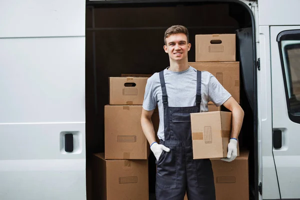 Un joven trabajador sonriente y guapo vestido de uniforme está de pie junto a la furgoneta llena de cajas con una caja en las manos. Movimiento de casa, servicio de mudanza — Foto de Stock