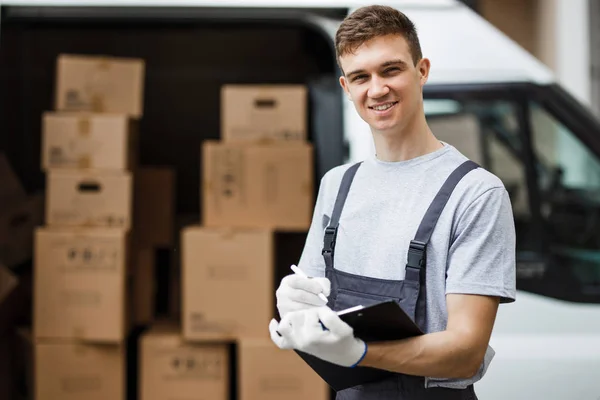 Młody przystojny pracownik uśmiechający się na sobie mundur stoi obok van pełne pudła przytrzymanie Schowka w jego ręce. Dom przesunąć, mover service. — Zdjęcie stockowe