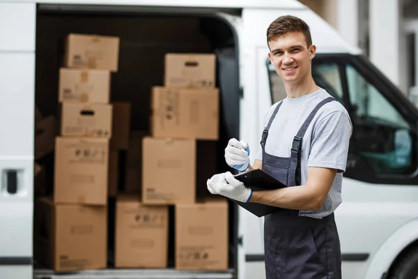 Un joven trabajador sonriente y guapo vestido de uniforme está de pie junto a la furgoneta llena de cajas con un portapapeles en las manos. Movimiento de casa, servicio de mudanza . — Foto de Stock