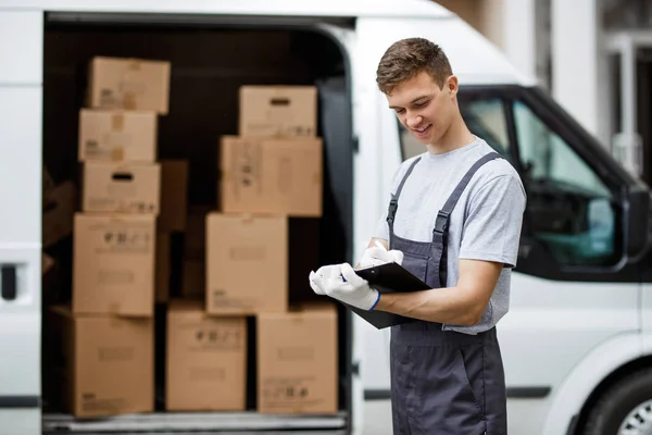 Un joven trabajador sonriente y guapo vestido de uniforme está de pie junto a la furgoneta llena de cajas con un portapapeles en las manos. Movimiento de casa, servicio de mudanza . — Foto de Stock