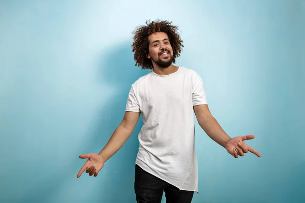 En curly-headed brunet man sträcker ut armarna med punkt fingrar uttrycker njutning. Asymmetrisk vit T-shirt över den blå bakgrunden. — Stockfoto