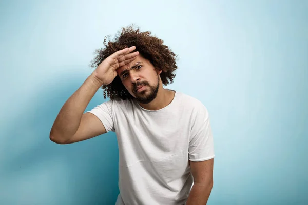En curly-headed brunet man håller hans palm över pannan ser koncentrerad och allvarliga. Kort skägg och tjocka ögonbryn. — Stockfoto