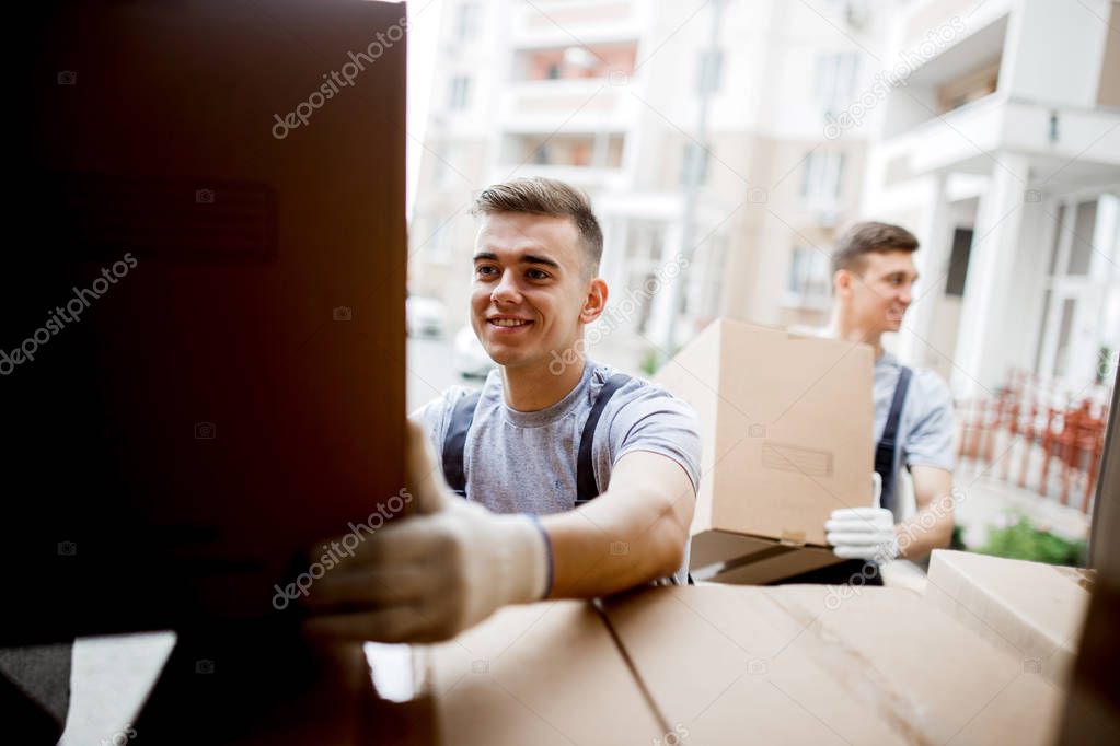 A young handsome smiling mover wearing uniform is reaching for the box while unloading the van full of boxes. House move, mover service