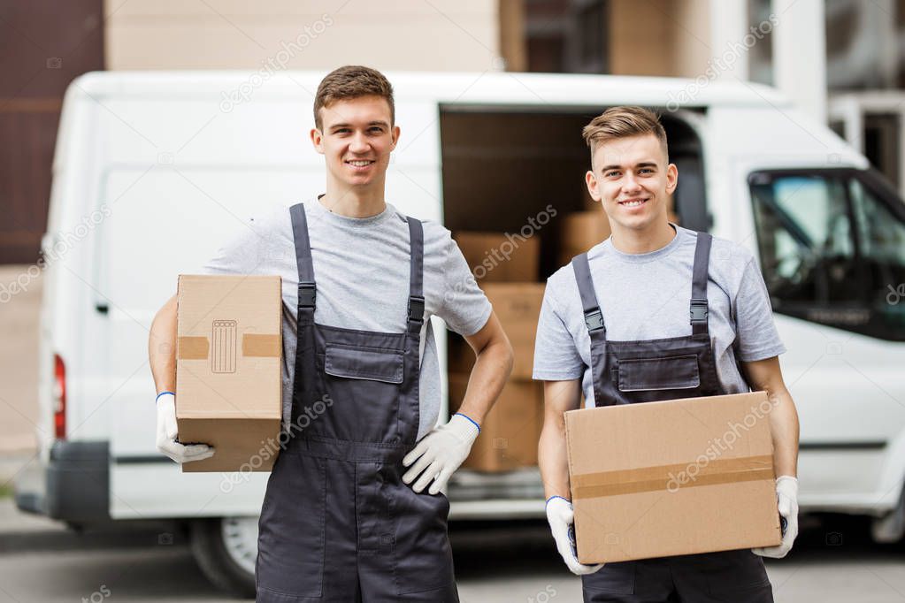 Two young handsome smiling workers wearing uniforms are standing in front of the van full of boxes holding boxes in their hands. House move, mover service