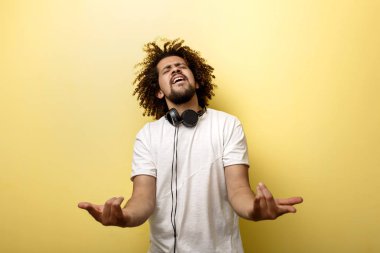 Bir bukle ve boyun kulaklık onun saçlı bir adam ellerini önünde tutuyor ve sarı arka plan üzerinde gözlerini tutmak bir boynuz jest yapma kapalı