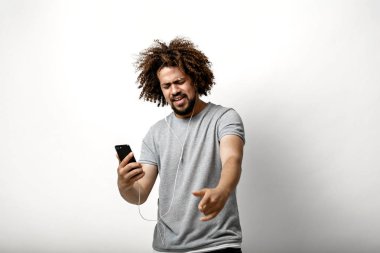 Bir kıvırcık kafalı yakışıklı adam uçan saç ve mutlu bir gülümseme ile gri bir T-shirt giyen kulaklık müzik dinleme ve beyaz arka plan üzerinde dans