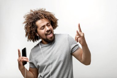 Gri bir T-shirt giyen kıvırcık kafalı yakışıklı bir adam mutlu bir gülümseme ile kulaklık müzik dinleme ve beyaz arka plan üzerinde boynuz jestler yapma