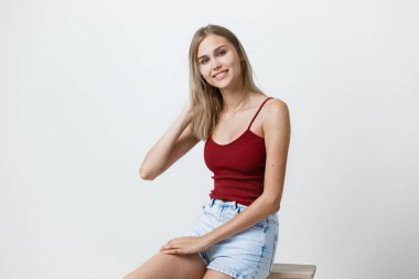 Bordo tişört ve mavi kot şort giyen, kamera, Studio ahşap sandalye üzerinde oturan bakarak gülen gülümseyen neşeli güzel genç sarışın kız