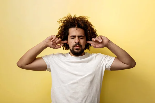 En curly-headed garvade man håller fingrarna i öronen, ögonen är stängda i ovilja att lyssna. Vit T-shirt över gul bakgrund — Stockfoto