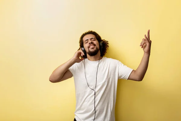 En curly-headed man klädd i vit T-shirt lyssnar på musik och göra ett horn gest med handen. Härlig stämning och bliss — Stockfoto