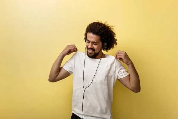 En curly-headed garvade man håller sin vita T-shirt med fingertopparna medan du lyssnar på musik i hörlurarna och leende — Stockfoto