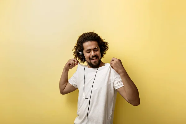 En curly-headed garvade man håller sin vita T-shirt med fingertopparna medan du lyssnar på musik i hörlurarna och charmigt leende — Stockfoto