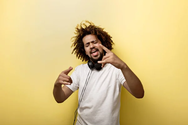 A man with his hair in a frizz and headphones on the neck is holding his hands in front of him and making a horn gesture over the yellow background