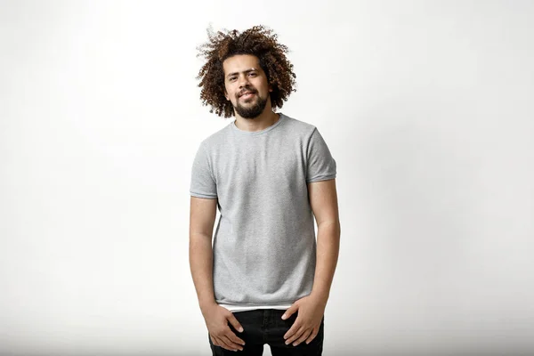 En curly-headed stilig man klädd i en grå T-shirt står något leende över den vita bakgrunden — Stockfoto