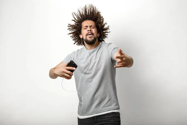 En curly-headed stilig man klädd i en grå T-shirt med flygande hår och ett lyckligt leende lyssnar på musik i hörlurarna och dansar över den vita bakgrunden — Stockfoto