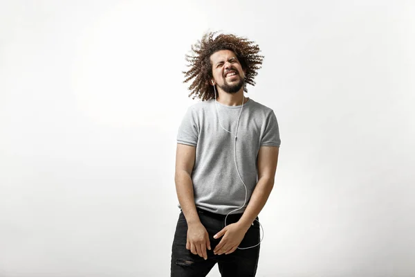 En curly-headed stilig man klädd i en grå T-shirt lyssnar på musik i hörlurar med ett glatt leende och flygande hår över den vita bakgrunden — Stockfoto