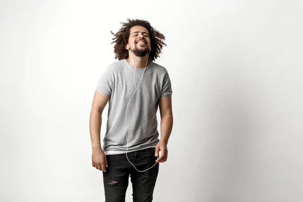 En curly-headed stilig man klädd i en grå T-shirt och slet jeans lyssnar på musik i hörlurarna med en lycksalig uttryck i ansiktet över den vita bakgrunden — Stockfoto