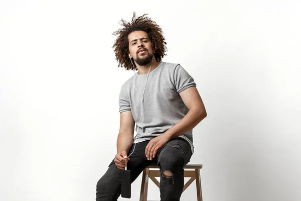 A curly-headed handsome man wearing a gray T-shirt and ripped jeans is listening to music in the earphones and sitting on the backless stool over the white background