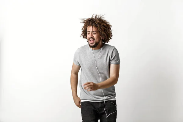 En curly-headed stilig man klädd i en grå T-shirt och slet jeans lyssnar på musik i hörlurarna och dansar över den vita bakgrunden — Stockfoto