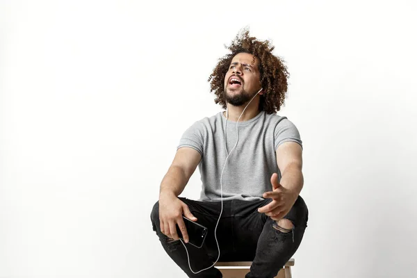 En curly-headed stilig man klädd i en grå T-shirt och slet jeans Lyssna på musik i hörlurar, sitter på den backless pallen och sjunger över den vita bakgrunden — Stockfoto