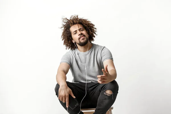 A curly-headed handsome man wearing a gray T-shirt and ripped jeans is listening to music in the earphones and sitting on the backless stool with a confident outlook over the white background