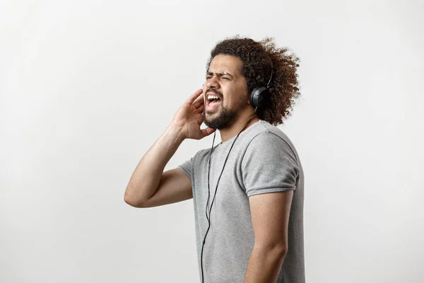 En curly-headed stilig man klädd i en grå T-shirt och slet jeans är ständiga och lyssnar på musik i hörlurarna och sjunger passionerat över den vita bakgrunden — Stockfoto