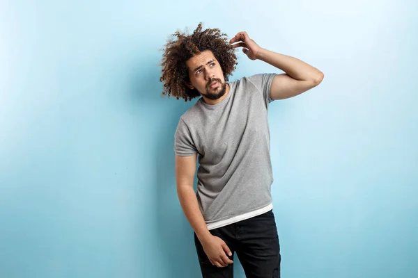 En curly-headed stilig man klädd i en grå T-shirt och slet jeans är stående och tänkande med handen upp och röra håret över den blå bakgrunden — Stockfoto