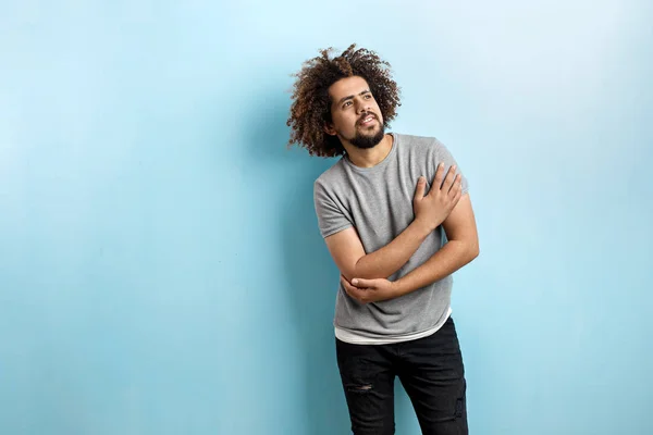 En curly-headed stilig man klädd i en grå T-shirt och slet jeans är stående och tänkande med handen på bröstet, titta uppåt över den blå bakgrunden — Stockfoto