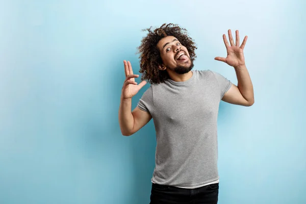 En curly-headed stilig man klädd i en grå T-shirt är att ha kul med tungan ut, lekfull ögon och handflatorna öppna och upp över den blå bakgrunden — Stockfoto