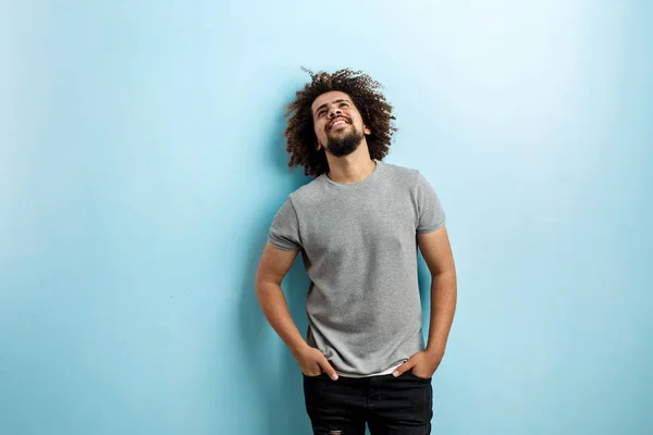 En curly-headed stilig man klädd i en grå T-shirt och slet jeans är stående och leende med händerna i fickorna, tittar uppåt över den blå bakgrunden — Stockfoto