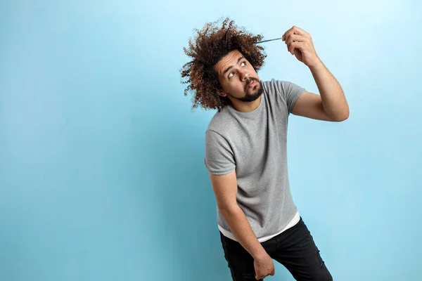 En curly-headed stilig man klädd i en grå T-shirt stående med en tankeväckande och koncentrerad blick och röra vid sitt hår över den blå bakgrunden — Stockfoto