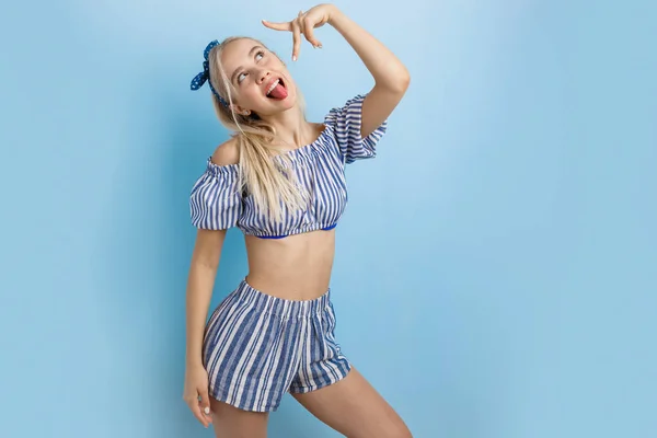 Menina loira bonita alegre e animado em roupa de verão, mostrando a língua, posando para a câmera, de pé sobre o fundo azul — Fotografia de Stock