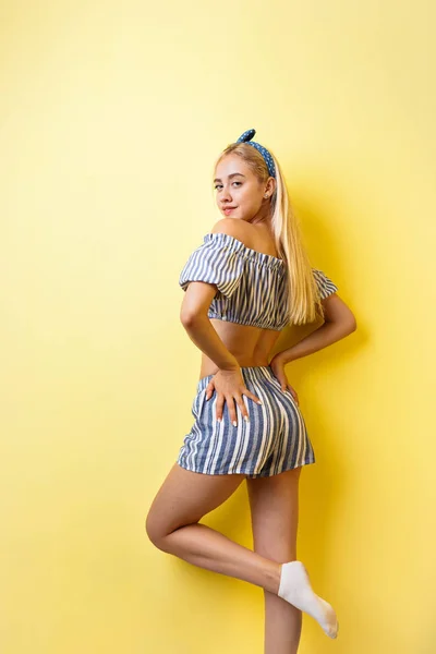 Joyful beautiful blonde girl in summer outfit is smiling standing in a half-turn, lifting one leg up, over yellow background. Emotions, gestures and facial expression