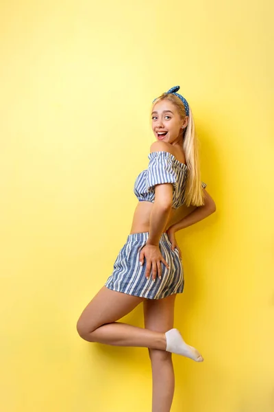 Joyful beautiful blonde girl in summer outfit is smiling standing in a half-turn, lifting one leg up, over yellow background. Emotions, gestures and facial expression