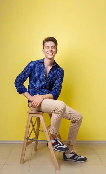 Knappe vriendelijke op zoek vrolijke Europese guy dragen blauw shirt en beige broek lachend zittend op houten kruk geel muur — Stockfoto
