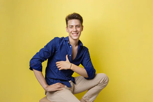 Bonito amigável olhando alegre europeu cara vestindo camisa azul e bege calças sorrindo enquanto sentado em banquinho de madeira sobre a parede amarela — Fotografia de Stock