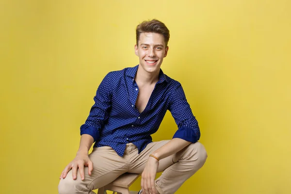 Krásný, přátelský, vesele Evropské chlap na sobě modrou košili a béžové kalhoty s úsměvem při posezení na dřevěné stolici nad žlutá stěna — Stock fotografie