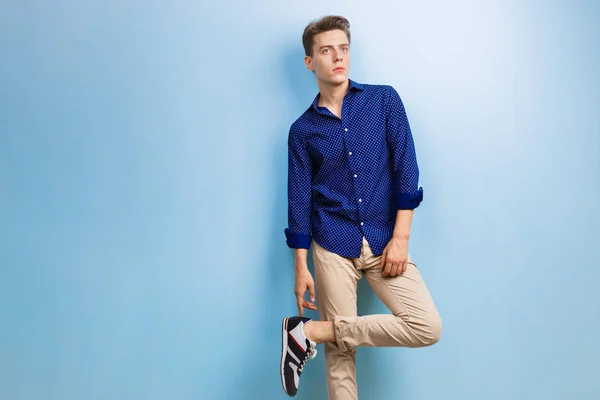 Calma riflessivo bel ragazzo dai capelli scuri indossa camicia blu e pantaloni beige, in piedi contro il muro blu — Foto Stock