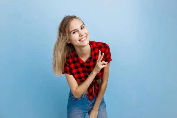 Vrolijke knap blond meisje met blauwe ogen het dragen van rode geruite shirt en spijkerbroek, v-teken, permanent tegen blauwe muur tonen — Stockfoto