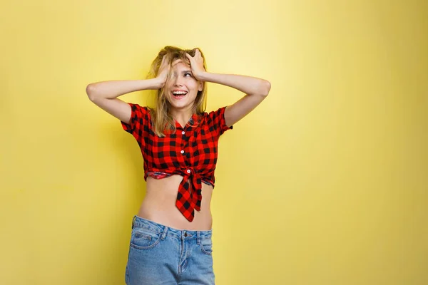 Vrolijke knap jong blond meisje met blauwe ogen het dragen van rode geruite shirt en spijkerbroek, met handen op haar hoofd, wordt opgewekt, permanent tegen gele muur — Stockfoto