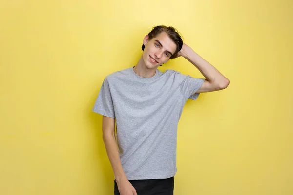 Glada stilig ung tunn mörkhårig kille med blå ögon bär grå t-shirt hålla handen på huvudet, stående mot gul bakgrund — Stockfoto